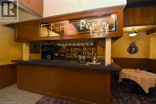 Elegant bar featuring warm wood cabinetry, illuminated shelving displaying a curated liquor collection, mirrored backlit signage, and tile flooring - 942-946 Barton Street E, Hamilton, ON 