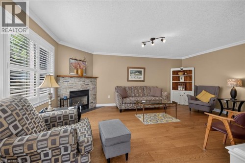 Living room with wood-finish flooring, stone-clad fireplace with wood mantel, crown molding, and window with plantation shutters - 10 Balvenie Boulevard, Caledonia, ON 