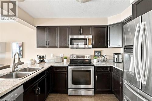 U-shaped kitchen featuring dark wood cabinetry, light-toned countertops, stainless steel appliances, and a double basin sink with a high-arc faucet - 187 Eden Oak Trail, Kitchener, ON 