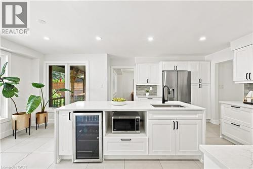 White shaker-style cabinetry with matte black hardware, stainless steel appliances, a built-in wine refrigerator, a large center island with an undermount sink, and light-toned tile flooring - 62 Corfield Drive, Kitchener, ON 