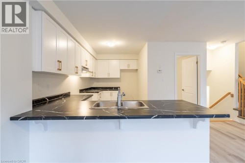 White cabinetry, dark stone-finish countertops, stainless steel sink, wood-finish flooring, and a natural wood staircase - 23 Dryden Lane, Hamilton, ON 