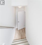 Carpeted staircase with a wood handrail, leading to a hallway with wood-finish flooring, a two-panel interior door, and a flush-mount ceiling light fixture - 