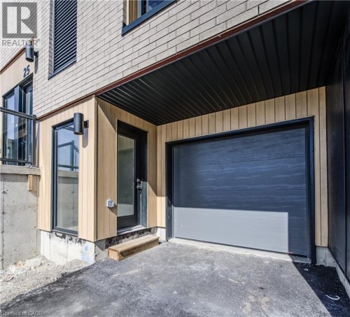 Exterior with light brick siding and wood-finish paneling, featuring a dark overhead garage door, a black framed entry door, and exterior wall sconces - 25 Urbane Boulevard, Kitchener, ON 