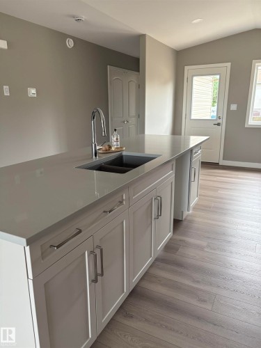 Kitchen featuring white cabinetry, light wood-style floors, a center island with sink, light stone counters, and lofted ceiling - 7413 44 Avenue, Camrose, AB - Indoor Photo Showing Kitchen With Double Sink