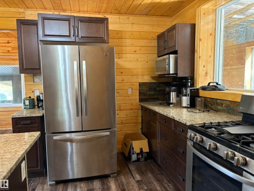 Kitchen with stainless steel appliances, dark brown cabinets, wooden walls, dark wood-type flooring, and light stone counters - B58018 Hiway 29, Rural St. Paul County, AB - Indoor Photo Showing Kitchen
