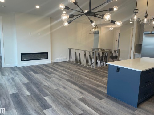 Two tone kitchen featuring hanging lights, dark wood-style floors, a glass covered fireplace, light stone counters, and a kitchen island - 4112 Ginsburg Place, Edmonton, AB - Indoor