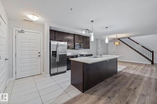 Kitchen with stainless steel appliances, dark brown cabinetry, an island with sink, decorative light fixtures, and light stone counters - 810 5151 Windermere Boulevard, Edmonton, AB - Indoor Photo Showing Kitchen With Stainless Steel Kitchen With Double Sink