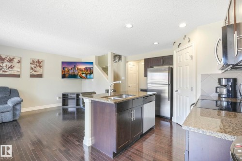 Kitchen featuring light stone countertops, dark brown cabinetry, appliances with stainless steel finishes, a textured ceiling, and a kitchen island with sink - 6819 Cardinal Link, Edmonton, AB - Indoor Photo Showing Kitchen With Double Sink
