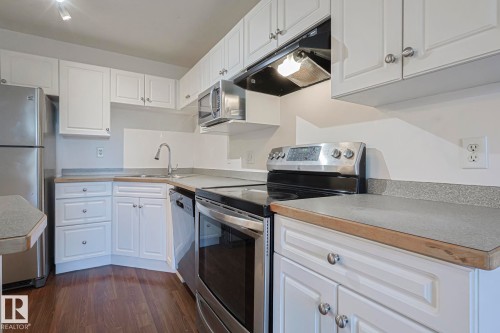 Kitchen featuring appliances with stainless steel finishes, white cabinetry, under cabinet range hood, dark wood finished floors, and light countertops - 120 7511 171 Street Nw, Edmonton, AB - Indoor Photo Showing Kitchen