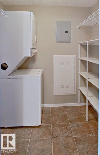 Laundry area with electric panel, stacked washer and clothes dryer, a textured ceiling, and light tile patterned floors - 404 5370 Chappelle Road, Edmonton, AB - Indoor