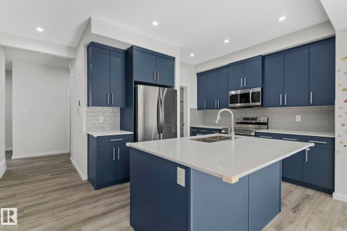 Kitchen with blue cabinets, a center island with sink, appliances with stainless steel finishes, light stone counters, and decorative backsplash - 1813 Collip View View, Edmonton, AB - Indoor Photo Showing Kitchen With Stainless Steel Kitchen With Double Sink With Upgraded Kitchen
