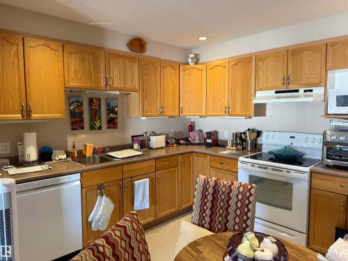 Kitchen featuring white appliances, a textured ceiling, under cabinet range hood, brown cabinets, and dark countertops - 415 4700 43 Avenue, Stony Plain, AB - Indoor Photo Showing Kitchen With Double Sink
