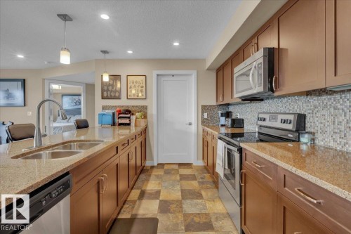 Kitchen featuring stainless steel appliances, light stone countertops, brown cabinets, a textured ceiling, and recessed lighting - 305 5029 Edgemont Boulevard, Edmonton, AB - Indoor Photo Showing Kitchen With Stainless Steel Kitchen With Double Sink With Upgraded Kitchen
