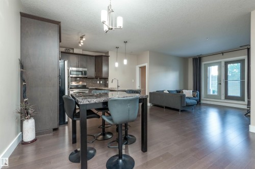 Kitchen featuring decorative backsplash, pendant lighting, light stone counters, dark wood-style flooring, and a textured ceiling - 140 6079 Maynard Way, Edmonton, AB - Indoor