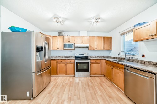 Kitchen with stainless steel appliances, light wood-style flooring, under cabinet range hood, a textured ceiling, and light brown cabinetry - #48 - 2503 24 Street Nw, Edmonton, AB - Indoor Photo Showing Kitchen With Double Sink