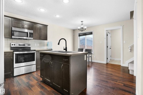 Kitchen with appliances with stainless steel finishes, dark brown cabinetry, a kitchen island with sink, decorative backsplash, and a chandelier - 1442 33A Street Nw, Edmonton, AB - Indoor Photo Showing Kitchen