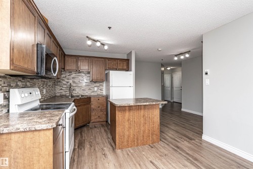 Kitchen featuring white appliances, rail lighting, a center island, backsplash, and a textured ceiling - 108 2305 35A Avenue, Edmonton, AB - Indoor Photo Showing Kitchen With Double Sink