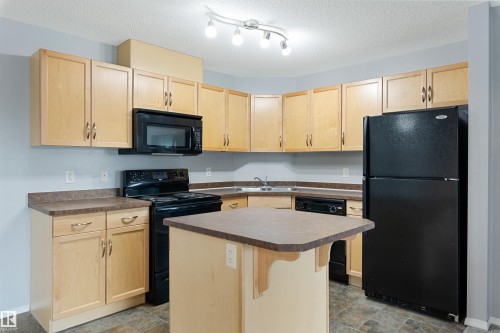 Kitchen featuring light brown cabinetry, black appliances, dark countertops, a center island, and a textured ceiling - 111 5951 165 Avenue, Edmonton, AB - Indoor Photo Showing Kitchen With Double Sink