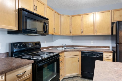 Kitchen with black appliances, light brown cabinetry, dark countertops, and a textured ceiling - 111 5951 165 Avenue, Edmonton, AB - Indoor Photo Showing Kitchen With Double Sink