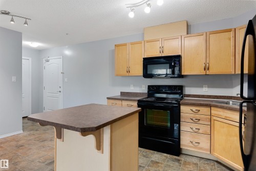 Kitchen with light brown cabinets, black appliances, a kitchen island, dark countertops, and track lighting - 111 5951 165 Avenue, Edmonton, AB - Indoor Photo Showing Kitchen