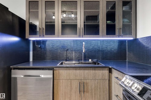 Kitchen with stainless steel appliances, light brown cabinetry, dark stone counters, and glass insert cabinets - 1702 10150 117 Street, Edmonton, AB - Indoor Photo Showing Kitchen With Double Sink
