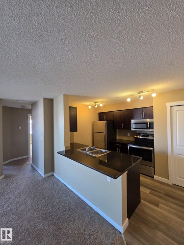 Kitchen with stainless steel appliances, dark brown cabinets, a peninsula, a textured ceiling, and a breakfast bar area - 313 1510 Watt Drive, Edmonton, AB - Indoor Photo Showing Kitchen With Double Sink