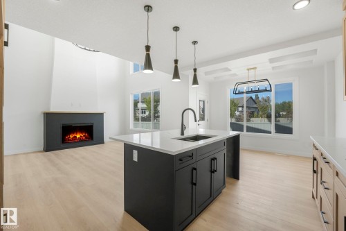 Kitchen featuring decorative light fixtures, dark cabinetry, light wood finished floors, a lit fireplace, and recessed lighting - 3140 Magpie Wy, Edmonton, AB - Indoor Photo Showing Kitchen With Fireplace