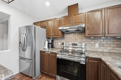 Kitchen with stainless steel appliances, under cabinet range hood, a textured ceiling, decorative backsplash, and brown cabinetry - 21017 60 Avenue, Edmonton, AB - Indoor Photo Showing Kitchen
