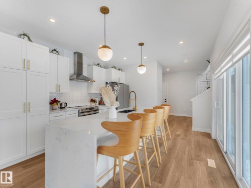 Kitchen featuring a breakfast bar area, decorative light fixtures, wall chimney range hood, light stone countertops, and backsplash - 4447 Kinsella Green, Edmonton, AB - Indoor Photo Showing Kitchen With Upgraded Kitchen