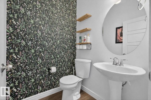 Bathroom featuring a pedestal sink with a chrome faucet, a large circular mirror, and patterned wallpaper - 8 Daniel Street, Fort Saskatchewan, AB - Indoor Photo Showing Bathroom