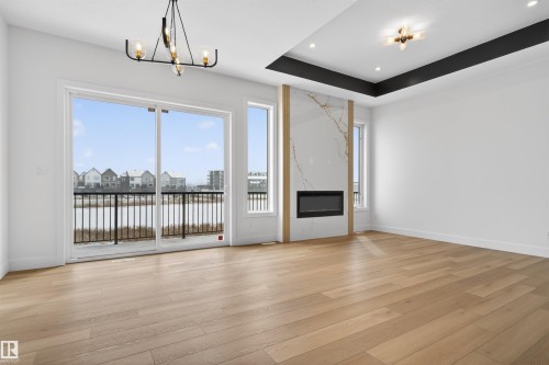 Spacious living area featuring light wood flooring, a decorative electric fireplace with a marble-style surround, and large sliding glass doors opening to a balcony with views of the water - 27 Rosewood Way, St. Albert, AB - Indoor With Fireplace