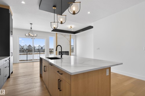 The property features an open-concept living area with light wood flooring, a kitchen island with a white countertop and dark sink, and large sliding glass doors providing views of a body of water and residences - 27 Rosewood Way, St. Albert, AB - Indoor Photo Showing Kitchen With Upgraded Kitchen