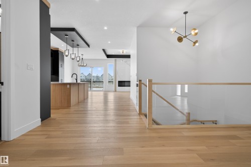 The property features light-toned hardwood flooring throughout, a staircase with a glass railing and wooden handrail, and a contemporary chandelier - 27 Rosewood Way, St. Albert, AB - Indoor Photo Showing Other Room