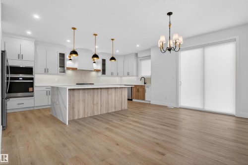 Open-concept kitchen featuring a large waterfall island with a wood-finish base, light wood-finish flooring, white cabinetry, stainless steel appliances, and a full-height tile backsplash - 27 Redpoll Wynd, St. Albert, AB - Indoor Photo Showing Kitchen With Upgraded Kitchen