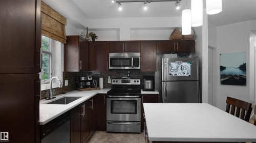Kitchen featuring stainless steel appliances, dark brown cabinets, a kitchen island, backsplash, and decorative light fixtures - 105 2584 Anderson Way, Edmonton, AB - Indoor Photo Showing Kitchen
