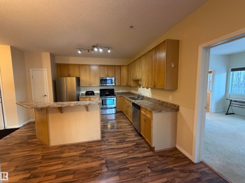 Kitchen featuring appliances with stainless steel finishes, a textured ceiling, dark wood-style floors, a breakfast bar area, and a kitchen island - 321 4304 139 Avenue, Edmonton, AB - Indoor Photo Showing Kitchen With Double Sink