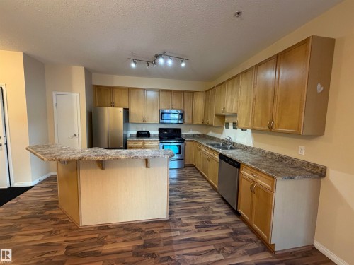 Kitchen with stainless steel appliances, a textured ceiling, a kitchen bar, dark wood-style flooring, and a kitchen island - 321 4304 139 Avenue, Edmonton, AB - Indoor Photo Showing Kitchen With Double Sink