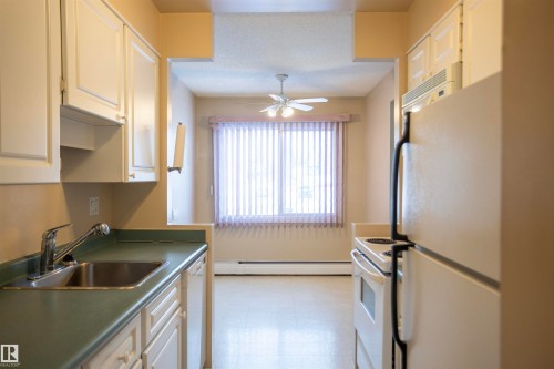 Kitchen with white appliances, light floors, a baseboard radiator, a ceiling fan, and white cabinetry - 207 11465 41 Avenue, Edmonton, AB - Indoor Photo Showing Kitchen