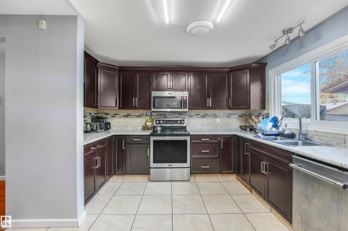 Kitchen with dark brown cabinetry, appliances with stainless steel finishes, light tile patterned floors, tasteful backsplash, and light stone counters - 13335 106 Street, Edmonton, AB - Indoor Photo Showing Kitchen With Double Sink