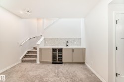 Indoor wet bar featuring beverage cooler, light carpet, light stone countertops, cream cabinets, and stairway - 