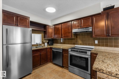 Kitchen featuring appliances with stainless steel finishes, backsplash, a textured ceiling, light stone counters, and under cabinet range hood - 289 Grandin Village, St. Albert, AB - Indoor Photo Showing Kitchen With Stainless Steel Kitchen With Double Sink