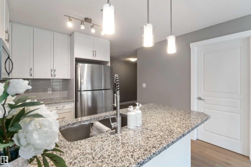 Kitchen with light stone counters, a peninsula, white cabinetry, stainless steel appliances, and hanging light fixtures - 101 3670 139 Avenue, Edmonton, AB - Indoor Photo Showing Kitchen With Upgraded Kitchen