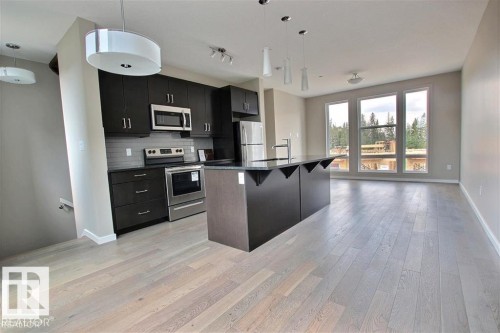 Kitchen with decorative light fixtures, stainless steel appliances, a kitchen breakfast bar, dark cabinetry, and a kitchen island with sink - 125 1304 Rutherford Road, Edmonton, AB - Indoor Photo Showing Kitchen With Upgraded Kitchen