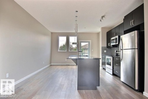 Kitchen featuring appliances with stainless steel finishes, dark cabinetry, a center island with sink, decorative light fixtures, and light wood-style flooring - 125 1304 Rutherford Road, Edmonton, AB - Indoor Photo Showing Kitchen