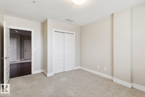 Unfurnished bedroom with light colored carpet, a closet, and a textured ceiling - 208 1350 Windermere Way, Edmonton, AB - Indoor Photo Showing Other Room