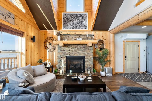 Living room featuring wood finished floors, a stone fireplace, wood walls, high vaulted ceiling, and a wall mounted air conditioner - 19003 Twp Rd 522, Rural Beaver County, AB - Indoor Photo Showing Living Room With Fireplace