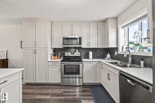 Kitchen with appliances with stainless steel finishes, a textured ceiling, backsplash, dark wood-type flooring, and light stone countertops - 20110 27 Avenue, Edmonton, AB - Indoor Photo Showing Kitchen With Double Sink