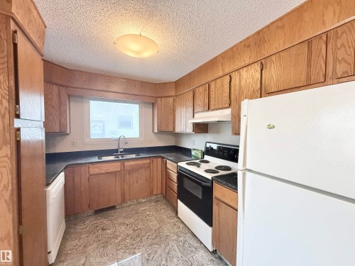 Kitchen with white appliances, dark countertops, a textured ceiling, brown cabinetry, and under cabinet range hood - 2112 104B Street, Edmonton, AB - Indoor Photo Showing Kitchen With Double Sink