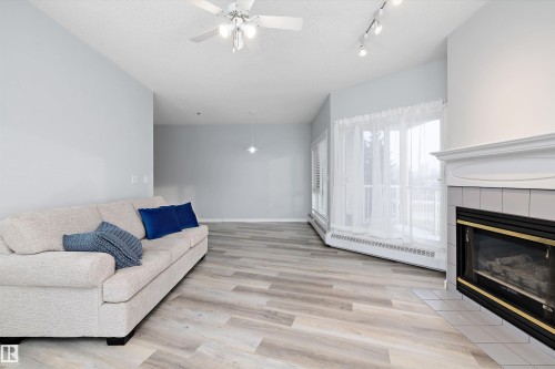 Living area with a textured ceiling, a ceiling fan, light wood-type flooring, a tile fireplace, and baseboard heating - 205 10809 Saskatchewan Drive, Edmonton, AB - Indoor Photo Showing Living Room With Fireplace