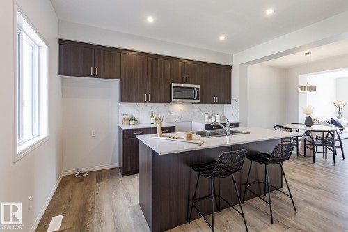 Kitchen with dark brown cabinetry, stainless steel microwave, light wood-style floors, an island with sink, and a breakfast bar - 3617 Triomphe Boulevard, Beaumont, AB - Indoor Photo Showing Kitchen With Double Sink With Upgraded Kitchen